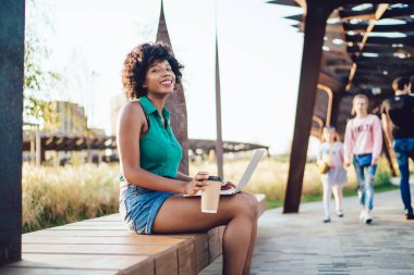 Mutlu, kıvırcık saçlı, yeşil tişörtlü, şortlu, dizüstü bilgisayarlı ve yazın arka planda insanlarla sokakta bir fincan kahveyle sörf yapan mutlu bir etnik kadın.