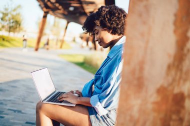 Gülümseyen kıvırcık saçlı, yeşil tişörtlü ve şortlu bir kadın yazın sokakta dizüstü bilgisayarda oturuyor. 