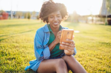 Afro saçlı, akıllı telefon kullanan ve yaz parkında çimlerde otururken kahve fincanıyla selfie çeken genç bir Afro-Amerikalı kadın.
