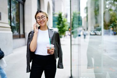 Deri ceketli, modern giyimli, gezici uluslararası iletişimin tadını çıkaran, gözlüklü mutlu Çinli kadın cep telefonu uygulaması ile konuşmak için telefon numarasını arıyor.