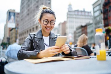 Klasik gözlüklü neşeli yeniyetme kız portresi. Elinde cep telefonu cihazları ve gülümseyen Asyalı blogcu sokak kafeteryasında oturmuş cep telefonlarını kullanmaktan zevk alıyor.