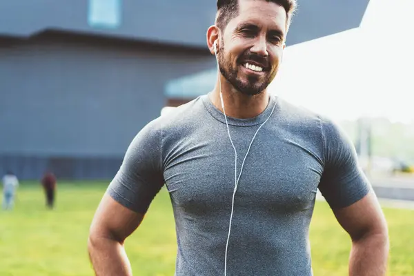 Happy smiling muscular man in sportswear with earphones listening to music preparing for sport exercise standing in street looking at camera