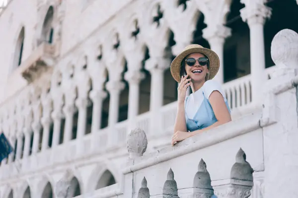 Cheerful Caucasian hipster girl using cheap tariffs in roaming making mobile phone call during journey on holidays, happy female tourist in hat enjoying wireless connection talking on cellular