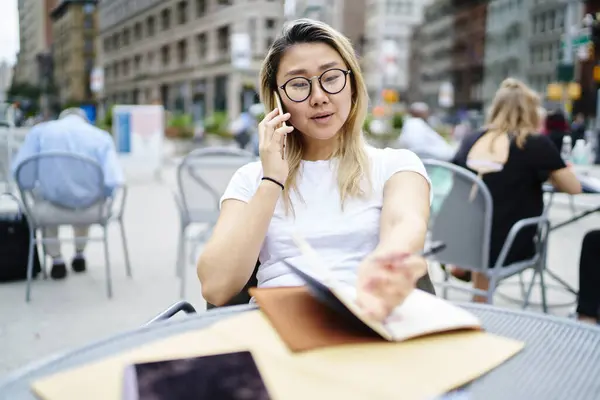 Chinese female millennial connecting to roaming while calling via cellphone application, Asian woman in glasses for vision protection making international phone call during leisure in street cafe