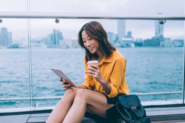 Hong Kong rıhtımında oturmuş kahve içerken sırt çantasıyla tablet kullanan sıradan tişörtlü ve etekli gülümseyen Asyalı genç kadının yan görüntüsü. 
