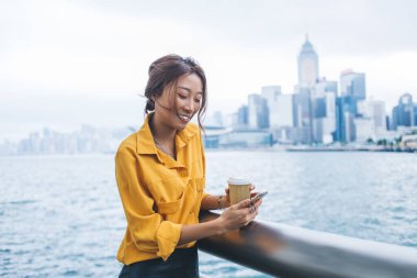 Elinde kahve fincanı tutan ve Hong Kong rıhtımında rüzgarlı bir günde korkuluklara tırmanırken akıllı telefonuyla mesajlaşan zarif, etnik bir kadın.