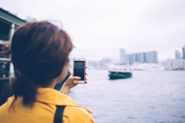 Sarı bluz giyen, Hong Kong körfezindeki yeşil feribotların ve modern gökdelenlerin dikey fotoğrafını beyaz cep telefonuyla çeken tanınmamış kadın turistin arka görüntüsü.