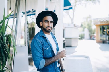 İspanyol, barışçıl, şık şapkalı, kot pantolonlu, şık şapkalı, şehirde kameraya bakmak için kahve taşıyan vahşi bir adam.