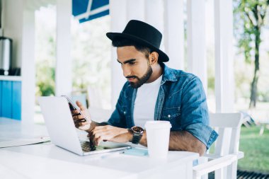 Şık, düşünceli, siyah şapkalı, günlük mavi ve beyaz tişörtlü, ekrana odaklanan ve laptopla etkileşime giren modern kafede yemek servisi yapan sıcak içeceklerle masada oturan ve akıllı telefon kullanan entelektüel bir adam.
