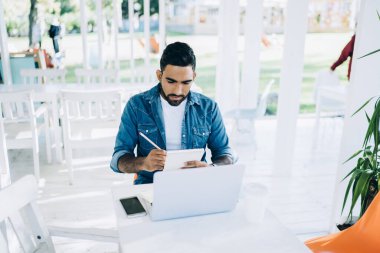Dışarıdaki kafenin terasında dizüstü bilgisayar incelerken günlük giysiler içinde notlar alan konsantre etnik erkek öğrencilerden. 