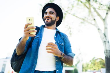 Gülümseyen, siyah şapkalı, beyaz t-shirt ve kot gömlekli, parkta cep telefonu kullanan ve yüzünü çeviren etnik bir erkek.