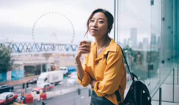 Half length portrait of pretty hipster girl smiling at camera during coffee time at urban setting, attractive female tourist with fashionable backpack holding takeaway cup enjoying holidays