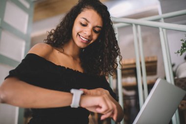 İyimser etnik bayan günlük kıyafetlerini giyiyor. Akıllı saate bakıyor. Kafedeki merdivenlerde oturan üniversite projesi için dizüstü bilgisayar kullanıyor.