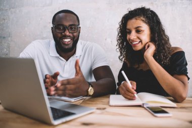 Neşeli Afro-Amerikalı erkek ve bayan meslektaşlar dizüstü bilgisayarda araştırma yaparak proje fikirlerini tartışıyor, koyu tenli adam Webinar aracılığıyla kız öğrenciye bilgiyi açıklarken gülümsüyor.