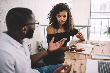 Kafası karışmış kaşları çatık kadın elinde kalemle dizüstü bilgisayarını işaret ederek kafede oturan Afro-Amerikan bir adamla proje stratejisini tartışıyor.