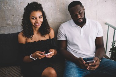 Kafedeki arkadaşıyla cep telefonuyla iletişim kurarken gülümseyen ve kameraya bakan siyah günlük giysili, tatmin olmuş Afrikalı Amerikalı bir kadından.