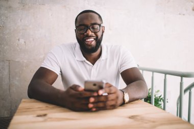 Beyaz tişörtlü ve gözlüklü, modern kafede akıllı telefondan iletişim kurarken gülen tatmin olmuş Afro-Amerikalı bir adam. 