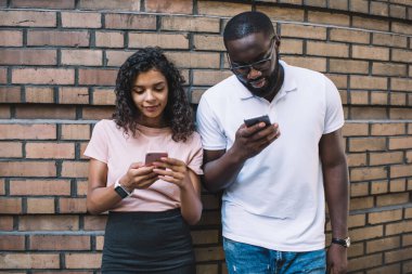 Koyu tenli genç erkek ve kadın milenyumlar sosyal ağlarda cep telefonlarına odaklanmış duvarın yanında birlikte duran Afro-Amerikan kadın ve erkek arkadaşları modern cihazlara bağımlı