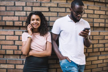 Neşeli Afrikalı Amerikalı hippi kız gezintide cep telefonuyla konuşuyor ve erkek arkadaşı cep telefonuyla mesajlar yolluyor. Koyu tenli erkek ve kadın arkadaşları sokak kullanım aletleri üzerinde birlikte duruyorlar.