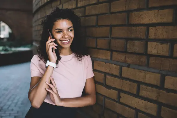 Cheerful curly dark skinned hipster girl standing on street satisfied with mobile phone connection in roaming, smiling charming african american 20s woman having conversation via cellular on street