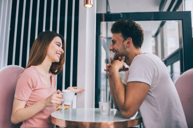 Çekici, mutlu kız arkadaşını dinleyen ve masada oturup kahve içerken gülen eli sıkı yakışıklı bir adamın yan görüntüsü.