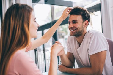 Neşeli, güler yüzlü, kafede eğlenen bir çiftin yan görünüşü. Kız arkadaşı partnerinin saçına dokunurken ve el ele tutuşurken.