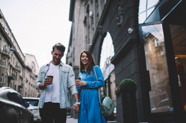 Aşağıdaki yakışıklı, memnun, sıradan kıyafetler giymiş, elinde içecekle kız arkadaşıyla el ele tutuşup gülen ve şehir merkezinde cadde boyunca yürüyen adamdan.