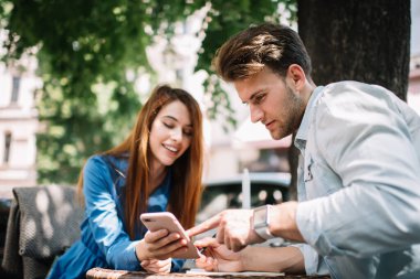 Akıllı telefon tutan neşeli, genç ve çekici bir kadın. Ve yakışıklı bir erkek arkadaşla bilgi paylaşıyor. Cihazı işaret ediyor.