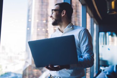 Resmi görevdeki iyimser gülümseyen adam, modern ofis alanının penceresinden bakarak başlangıç gelirini kontrol etmek için dizüstü bilgisayar kullanıyor.