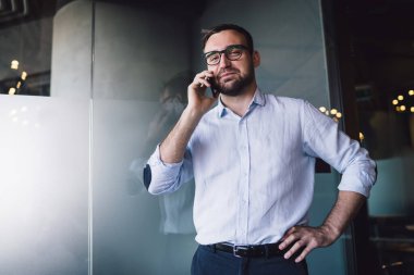 Mavi gömlekli ve mavi pantolonlu kendinden emin işadamı cam kapının karşısındaki ofiste cep telefonundan arama yapıyor.