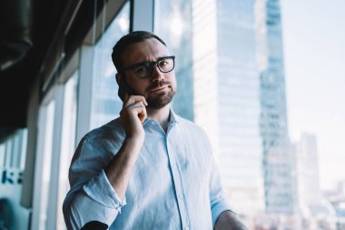 Gözlüklü, sakallı, ofisteki büyük pencerenin yanında dikilip kasvetli bir şekilde kameraya bakarken cep telefonuyla konuşan genç, tatminsiz yönetici erkek.