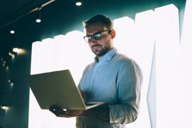 Gözlüklü, beyaz tişörtlü, aydınlatmalarla dekore edilmiş duvarın karşısında duran, dizüstü bilgisayarlı ciddi derecede konsantre olmuş bir girişimci.