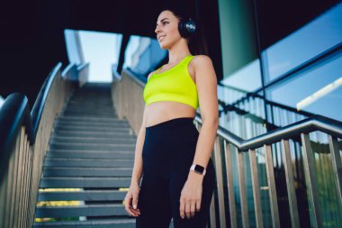 Spor kıyafetleriyle gülümseyen kadın yüzü ve güneşli bir günde modern binanın merdivenlerinde duran kulaklıklar.