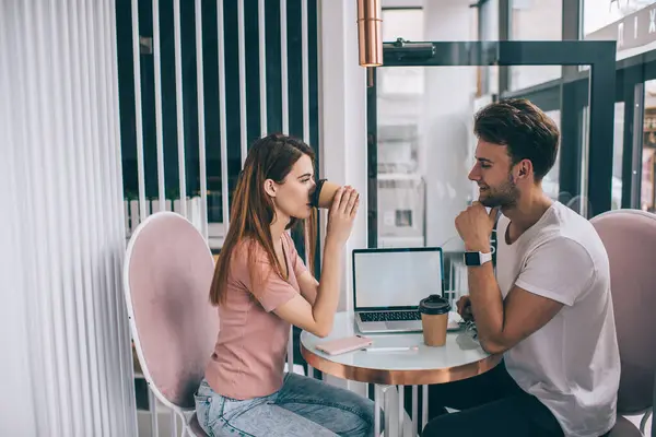 Thoughtful woman taking break from work and having coffee while meeting  talking and drinking cup of coffee with colleague with watch in cafeteria in sunlight