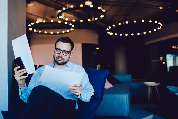 Молодой бизнесмен с щетиной и очками сидит в кресле со скрещенными ногами и внимательно анализирует бумаги со смартфоном в руках в современном офисе