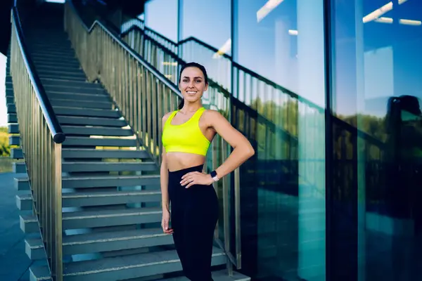 Half length portrait of happy female athlete in acid yellow top standing at urbanity and smiling at camera satisfied with healthy lifestyle, cheerful slim woman with perfect body shape posing