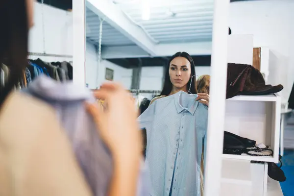 Stock image Caucasian woman with hanger trying shirt near mirror in merchandise boutique mall, attractive hipster girl posing near glass looking at own reflection while choosing brand clothing on shopping