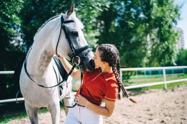 Güzel esmer profesyonel kadın jokey aygır ve öpüşen atın yanında duruyor, kadın binici çiftlikte tatildeyken hayvan arkadaşına bakmaya ve onunla ilgilenmeye hazırlanıyor.