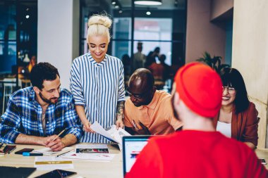 Gündelik kıyafetlerle doküman okuyup, masa başında birlikte çalışırken evrak işleriyle uğraşan bir grup genç çok ırklı iş arkadaşı.
