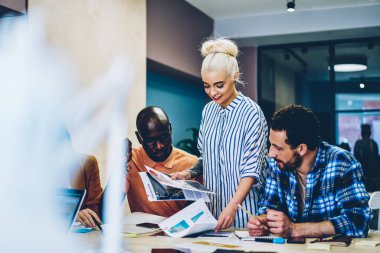 Günlük giysiler içinde kadın işçi çok ırklı iş arkadaşlarıyla kağıtları paylaşıyor ve çalışma alanında toplanırken başlangıç fikirlerini tartışıyorlar.