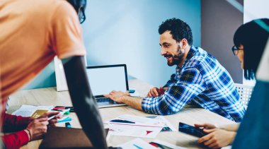 Üzerinde ekose gömlekli, boş ekranla yazı yazan ve gelecekte yapılacak işler için strateji geliştirirken çağdaş ofisteki çok ırklı iş ortaklarına görüşlerini açıklayan düşünceli yetişkinlerden.