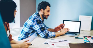 Gündelik giysiler içinde kendine güvenen yetişkin bir erkeğin boş ekran ile netbook kullanırken aynı zamanda ışık çağındaki ofisteki dalgın ekin meslektaşlarına görüşlerini açıklarken yan görüntüsü