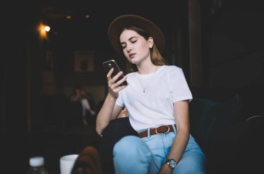 Ciddi bir beyaz kadın milenyumu modaya uygun giyim ve şapka cep telefonu uygulaması üzerinden kitap okumaya odaklı, düşünceli hippi kız akıllı telefon ve 4G kullanarak blogda medya dosyalarını paylaşıyor