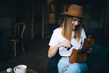 Moda giysiler içinde gülümseyen Kafkasyalı hippi kız ve kafenin içinde kredi kartı harcamak için debriyajını açan şapkası, şık havalı kıyafetleri olan neşeli 20 'li yaşlarda bir kadın çantasını kontrol ediyor.