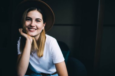 Moda şapkalı, siyah arka planda oturan neşeli 20 'li yaşlardaki hippi kızın yarım boy portresi. Gülümsüyor, şık giyinmiş, kameraya bakarak mutlu, genç bir kadın.