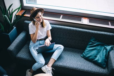 Moda şapkalı neşeli beyaz kadın, kafenin içinde koltukta oturuyor. Pozitif cep telefonu sohbetinden memnun, gülümseyen kadın servis operatörüne akıllı telefon ediyor.