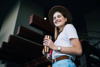 Şapkalı, şapkalı, gülümseyen, boş zamanın tadını çıkaran neşeli beyaz kadın portresi kitap ve içecekle termo bardak taşıyan zengin genç hippi kız.