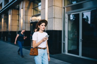 Moda kıyafetli çekici bir kadın. Arka planda geziyor. Cep telefonu taşıyor. Beyaz tenli, şık bir şapkayla şehir merkezine geliyor.