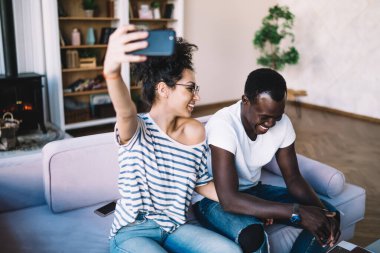 Tişörtlü, kot pantolonlu, akıllı telefondan selfie çeken ve gülen neşeli, çok ırklı bir çift.