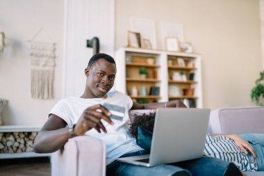 Afro-Amerikalı ciddi bir adam kameraya gülümseyerek bakıyor ve diz üstü bilgisayarlı bir kredi kartı tutuyor. Bu arada yan koltukta uyuyan etnik bir kadın var.